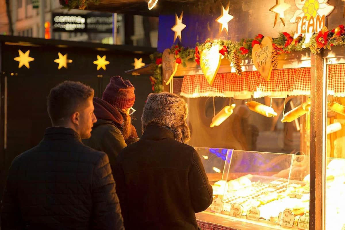 Vienna: Guided Schönbrunn Palace Christmas Market Tour + Apple Strudel Tasting Three people in winter clothing browse a festive outdoor market stall with decorative lights and holiday-themed decor.