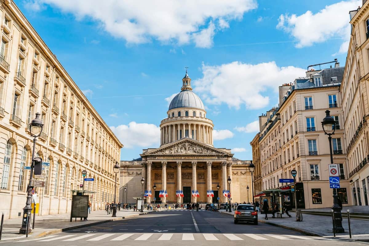 Paris : Visite guidée du Quartier Latin + Entrée au Panthéon Un bâtiment néoclassique avec des colonnes et un dôme, flanqué de bâtiments symétriques, vu d'un passage piéton sous un ciel partiellement nuageux.