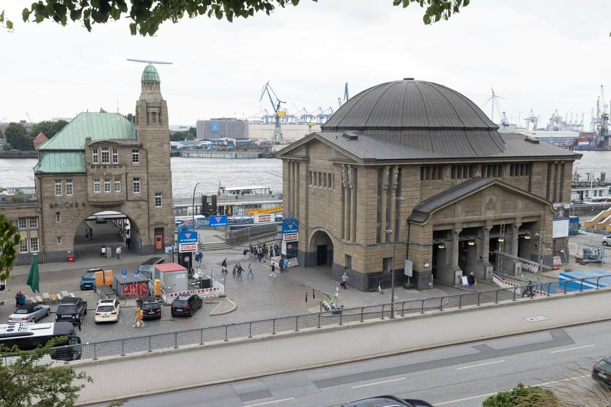 Hamburg: Riverfront Guided Walking Tour in German An urban scene featuring a large stone building by the waterfront, with cranes and industrial structures in the background. People and cars are visible.