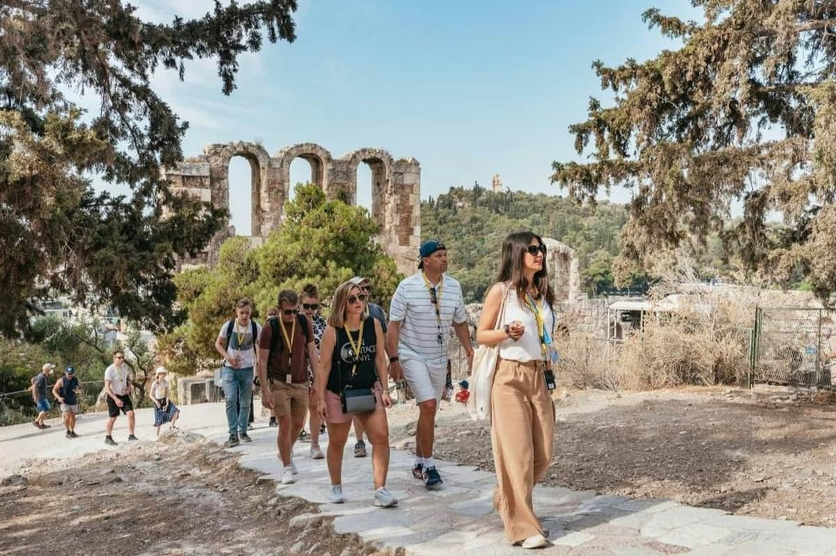 Acropolis & Ancient Agora of Athens: Skip The Line Ticket + Guided Tour A group of tourists walking on a stone path near ancient ruins surrounded by trees on a sunny day.