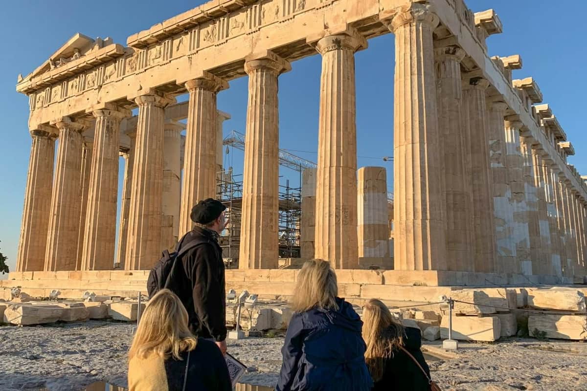La colline de l'Acropole et le musée de l'Acropole : visite guidée uniquement Four people observe the Parthenon, an ancient Greek temple with tall columns, under a clear blue sky. Scaffolding is visible inside.