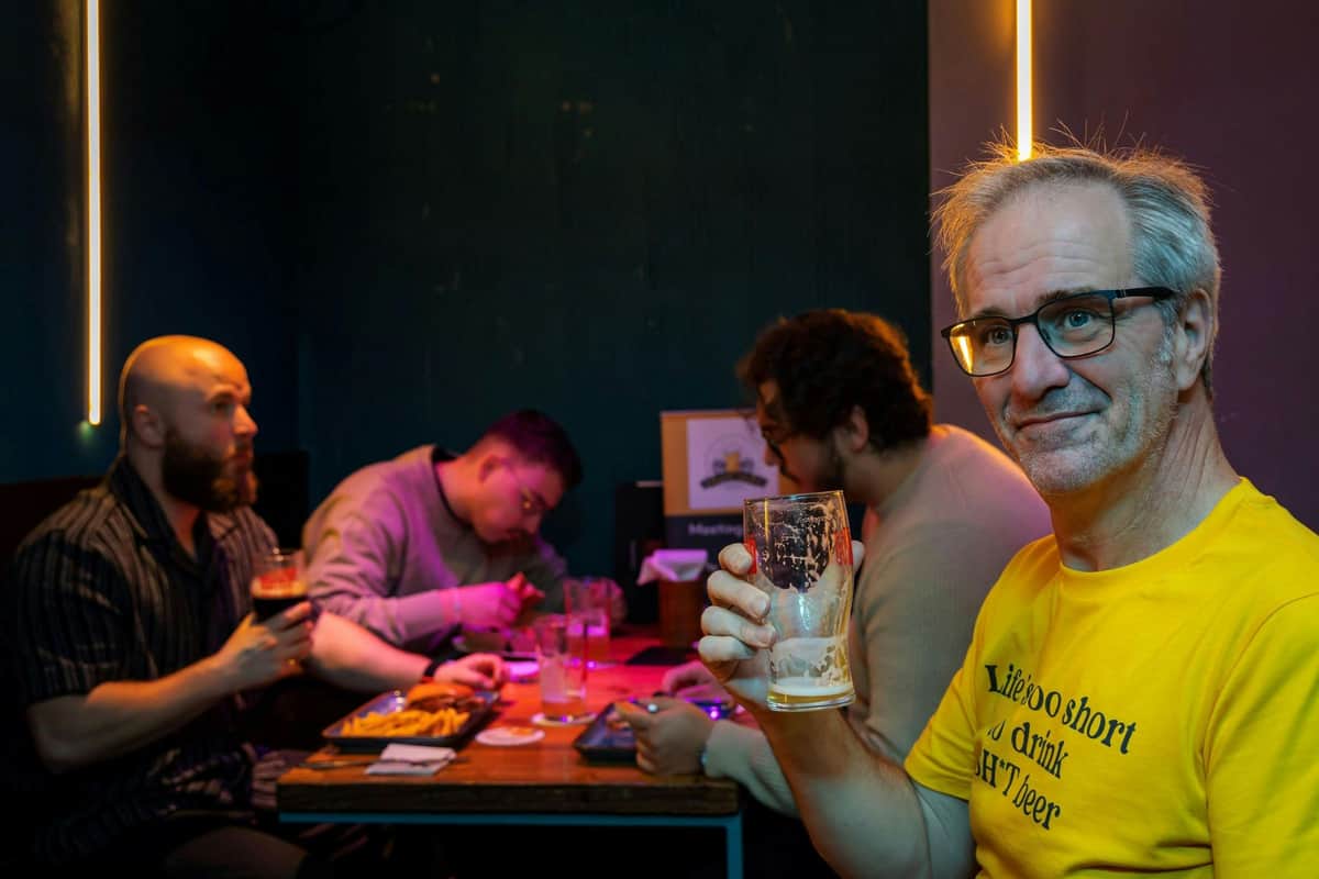 A man in a yellow shirt holding a glass in a dimly lit room with people sitting at a table in the background.