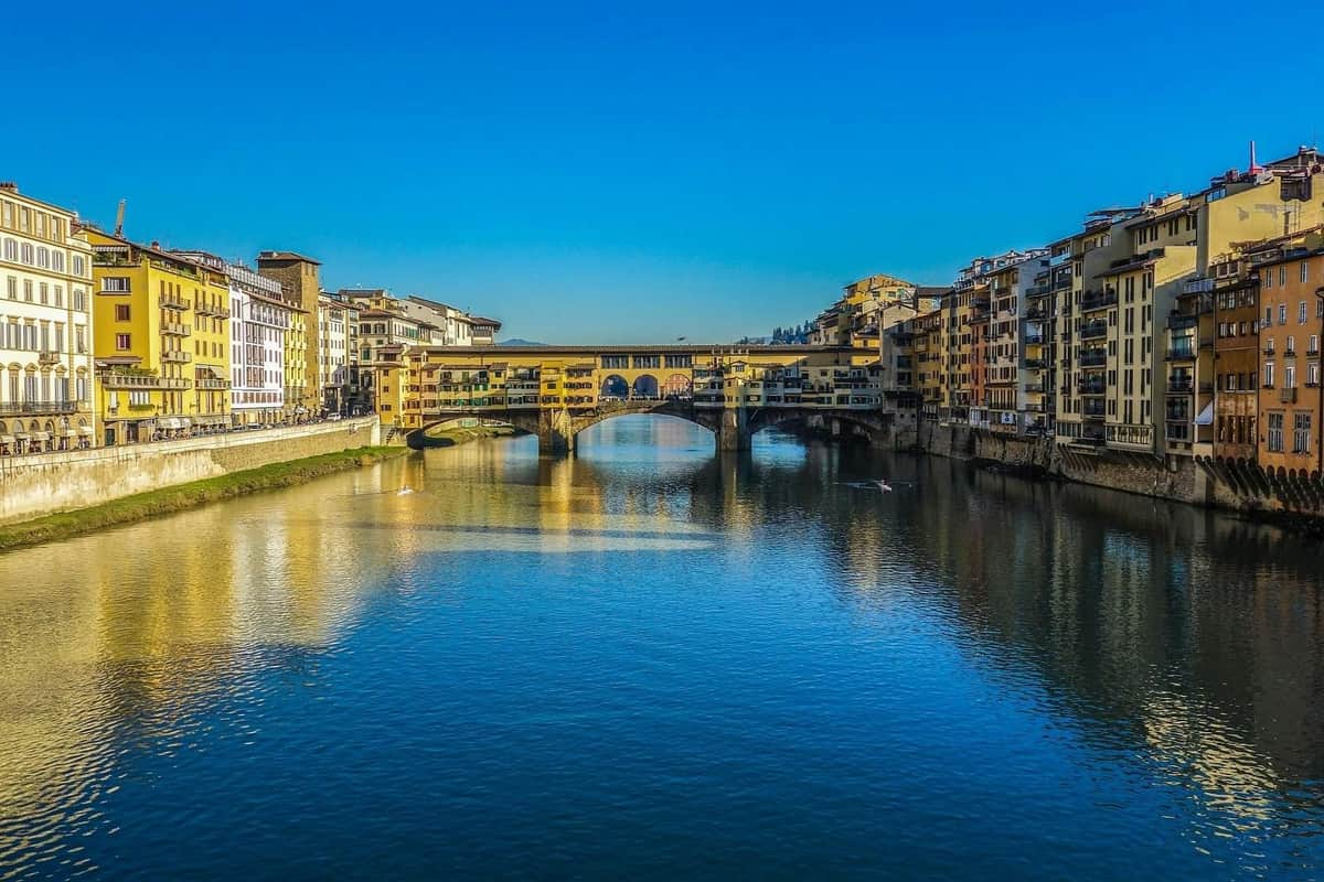 Uffizi Gallery & Vasari Corridor: Reserved Entry + Guided Tour A historic bridge over a calm river with buildings on both sides under a clear blue sky.