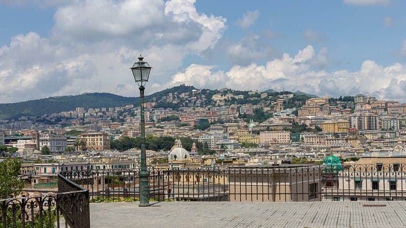 An ornate lamppost stands on a terrace overlooking a cityscape with hills, buildings, and a partly cloudy sky.