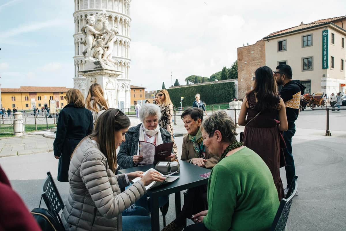 Florence and Pisa Shore Excursion from La Spezia: Priority Bus Access (La Spezia) People sitting at an outdoor café table near the Leaning Tower of Pisa, with others walking and interacting in the background.