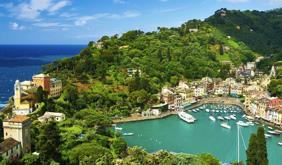 Genoa & Portofino: Daytrip from Milan A coastal town with colorful buildings, yachts docked in a turquoise harbor, surrounded by lush green hills and blue sky.