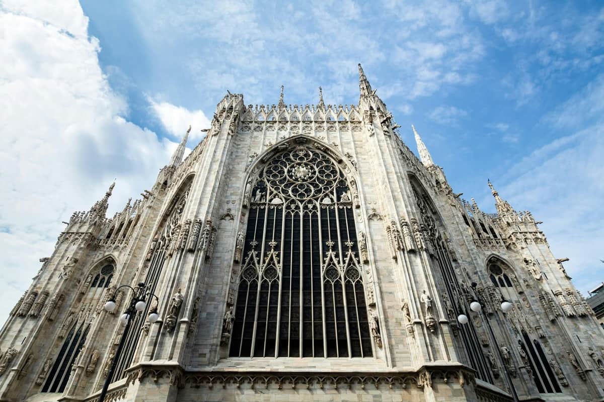 Milan : Visite guidée de la ville + Visite de la Cène et du Duomo Une façade de cathédrale gothique détaillée avec des sculptures complexes et des arcs en ogive, sous un ciel bleu partiellement nuageux.