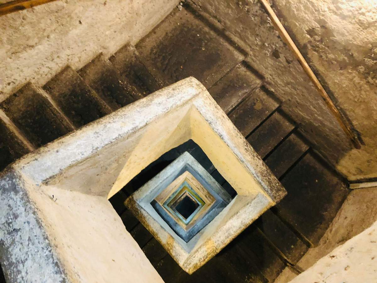 Vue aérienne d'un escalier en colimaçon carré en béton patiné. Les marches descendent en serpentant selon un motif géométrique.