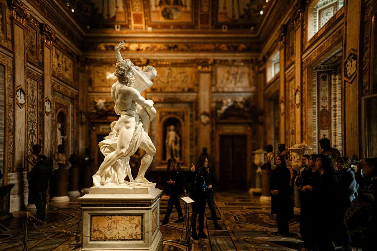 Borghese Gallery: Semi-Private Guided Tour A museum gallery featuring a marble statue prominently displayed in the center, with visitors observing the artwork.