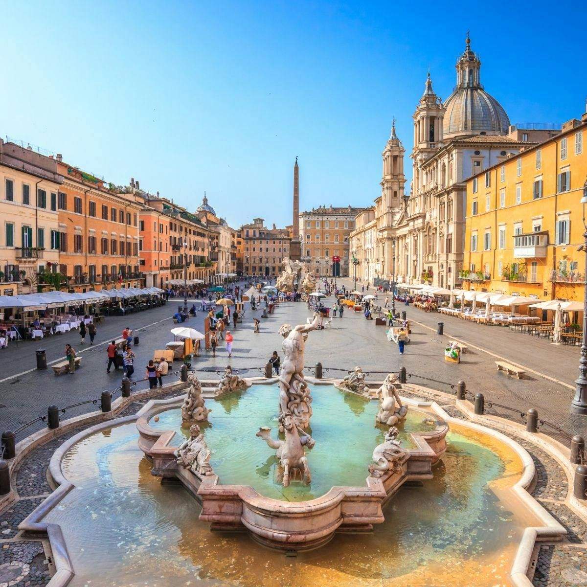 Piazza Navona, Pantheon, Trevi Fountain & Spanish Steps: Guided Walking Tour A lively square with a central fountain, surrounded by historic buildings, cafes, and people walking and sitting.