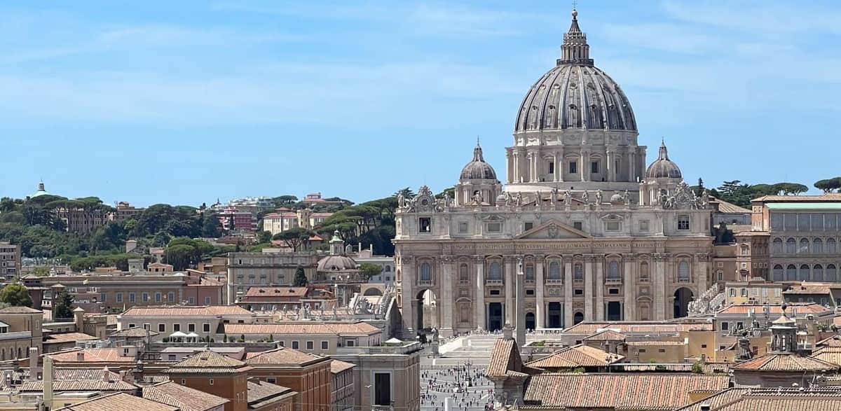 Basilique Saint-Pierre : Visite guidée + audience papale (Rome) Vue de la basilique Saint-Pierre avec son grand dôme, entourée de toits et d'arbres sous un ciel bleu clair.