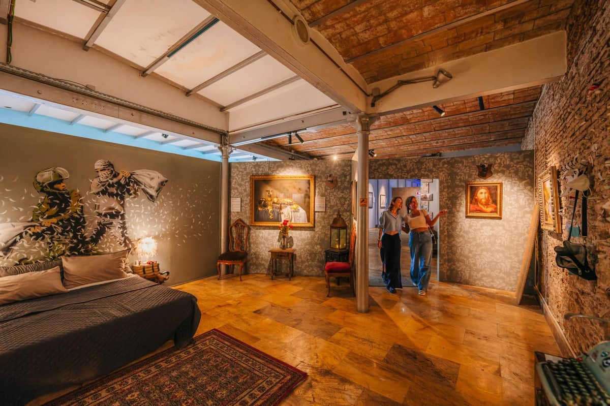 Banksy Museum Barcelona: Guided Tour Two people walk through an art-adorned room, featuring a bed, chairs, wall art, and exposed brick ceiling.