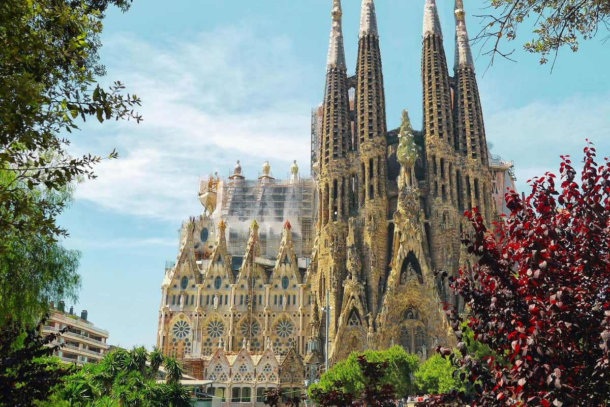 A large, intricate Gothic-style cathedral with multiple spires under a blue sky, surrounded by greenery and trees.