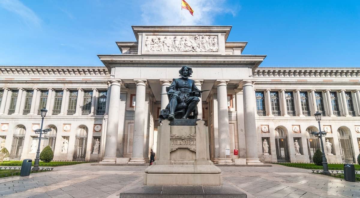 Musée du Prado et Palais Royal de Madrid : coupe-file et visite guidée (Madrid) Statue de Diego Velázquez devant les colonnes du musée du Prado, avec un drapeau espagnol flottant au-dessus.
