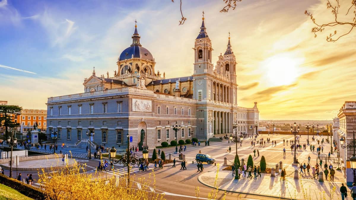 Un grand bâtiment avec des dômes et des tours sous un ciel couchant, entouré de gens, de voitures et de jardins entretenus.