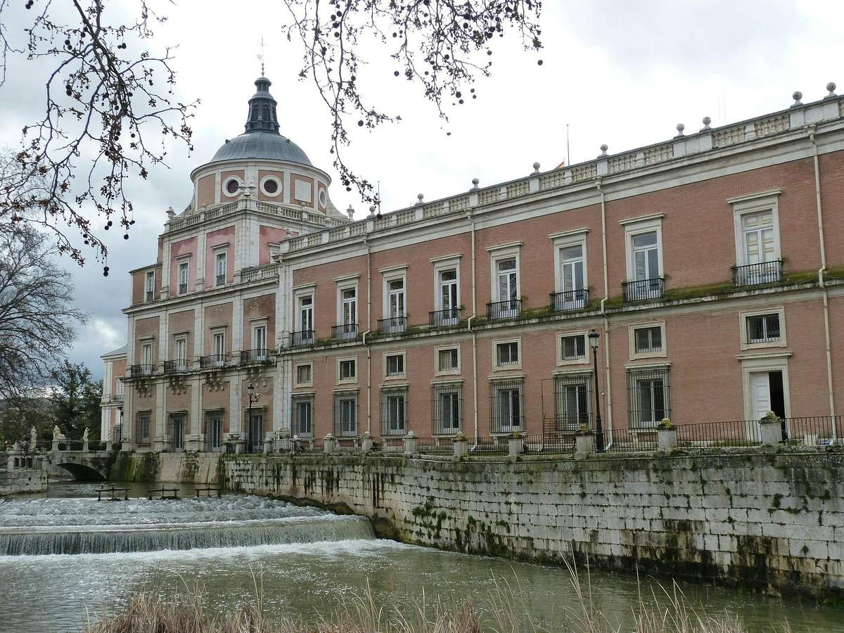 Palacio Real de Aranjuez: Führung (Madrid) Historisches Gebäude mit einer Kuppel, rosafarbener Backsteinfassade und zahlreichen Fenstern, direkt an einem reißenden Fluss unter einem bewölkten Himmel gelegen.