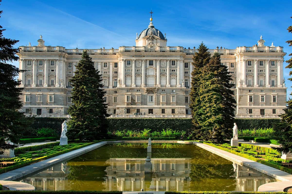 Palais royal de Madrid : Visite guidée (Madrid) Un palais majestueux avec une tour en forme de dôme, flanqué d'arbres, se reflétant dans un étang rectangulaire avec des statues dans un jardin formel.