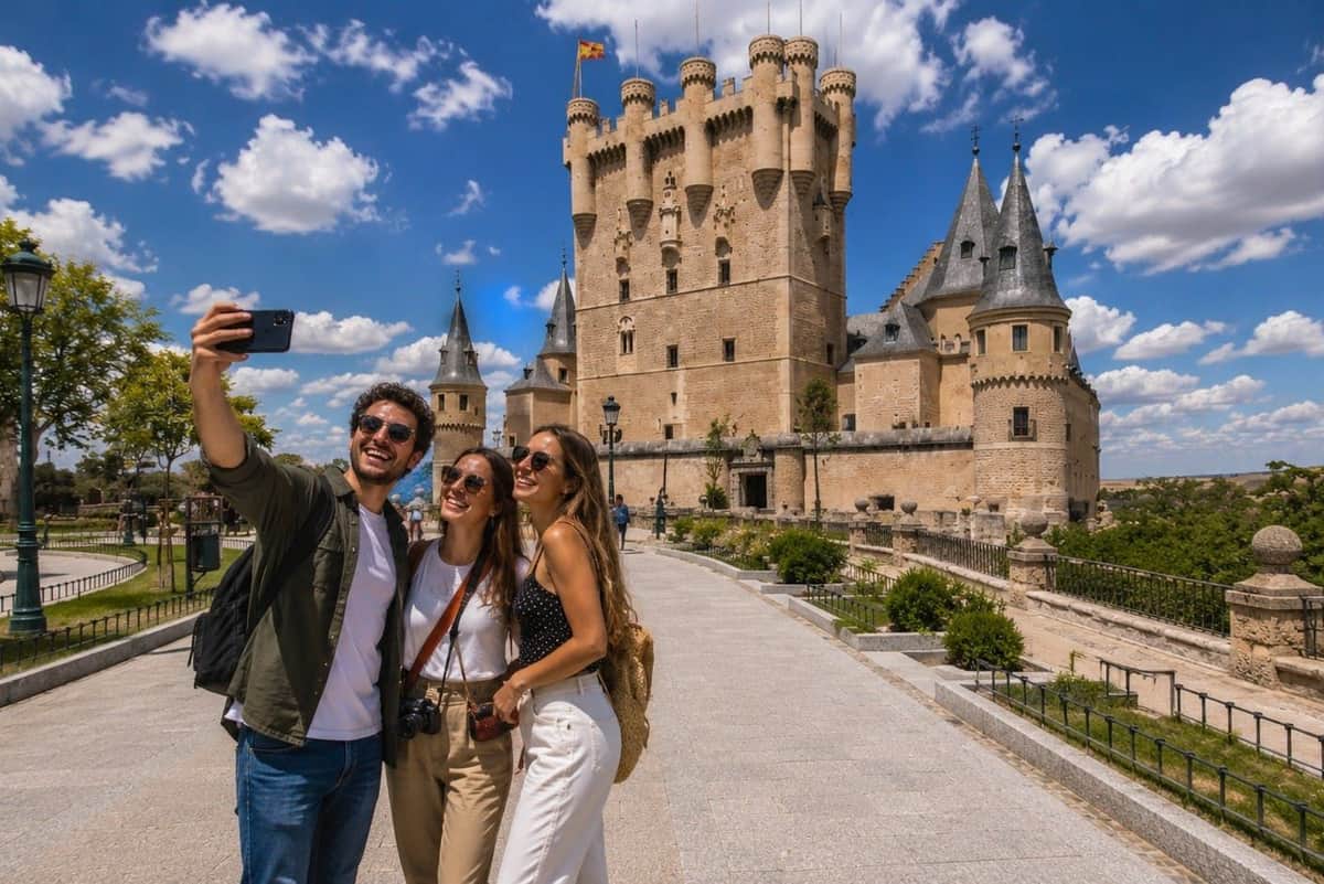 Ségovie et Avila : visite guidée à pied depuis Madrid Trois personnes sourient pour un selfie devant un château historique sous un ciel ensoleillé et nuageux.