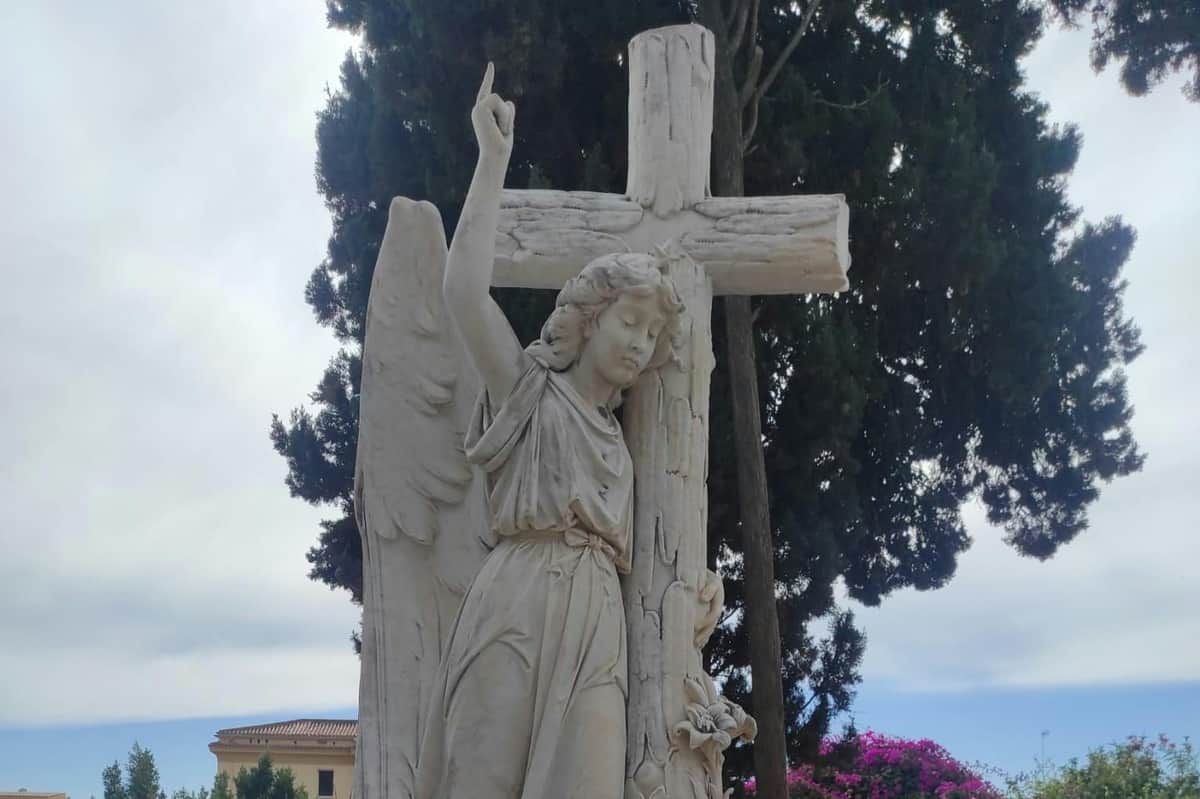 Malaga English Cemetery: Guided Tour (Málaga) A stone angel statue standing next to a cross, with trees and a building in the background.