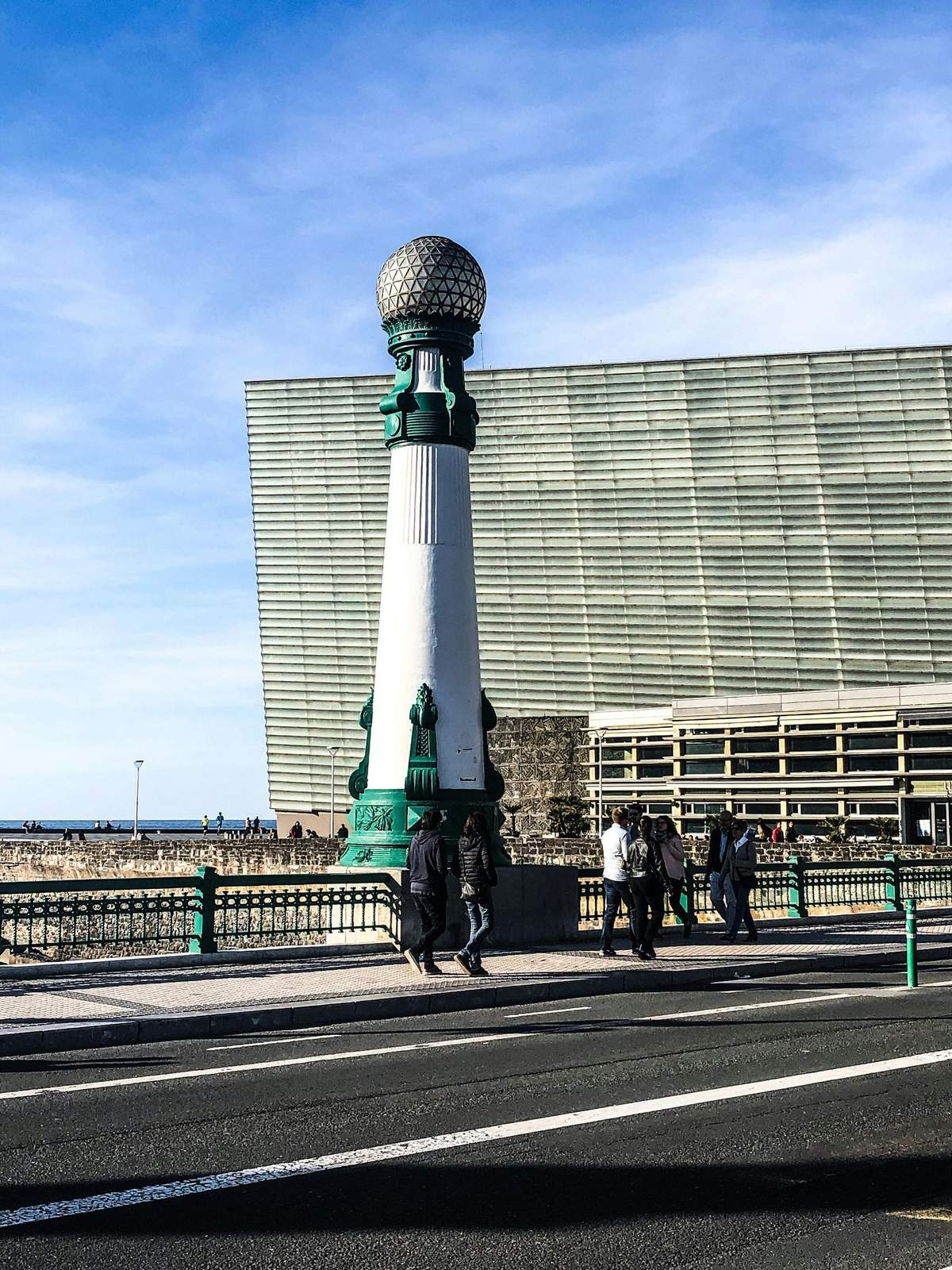 San Sebastián Historic & Cultural Walking Tour (San Sebastián) People walk by a tall, ornate green and white streetlight on a bridge, with a large modern building and the ocean in the background.