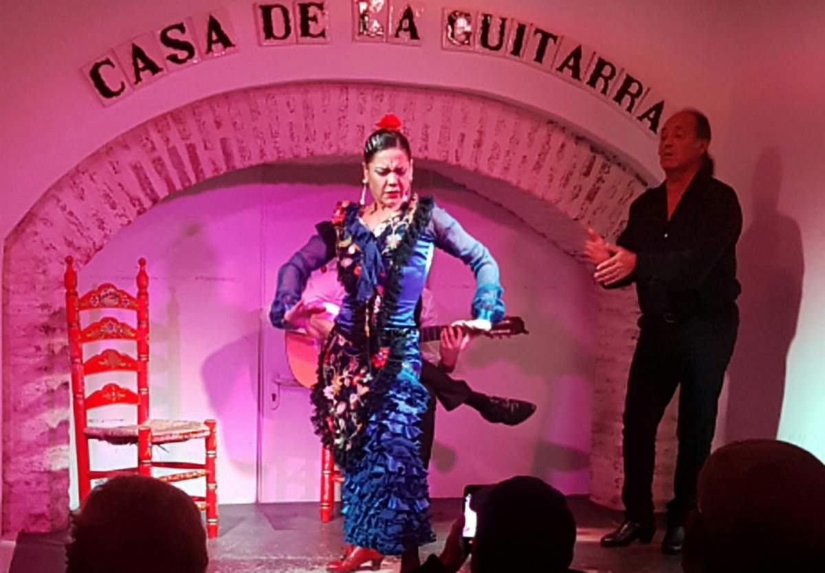 A flamenco dancer in a blue dress performs on a stage under an archway with "Casa de la Guitarra" sign, with a guitarist and chairs in the background.