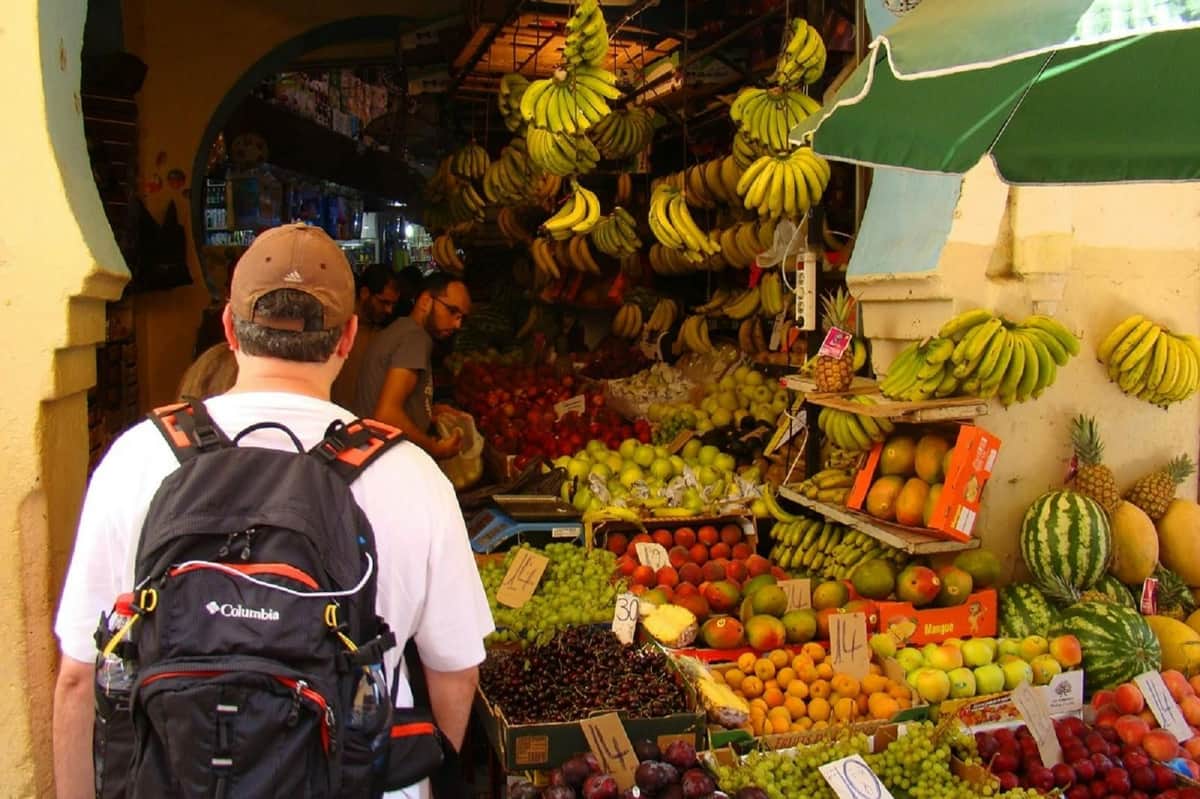 Tangier: Guided Day Trip from Seville + Ferry Ride + Lunch A man with a backpack browses a colorful fruit market, with hanging bananas and various fruits like apples, grapes, and mangoes.