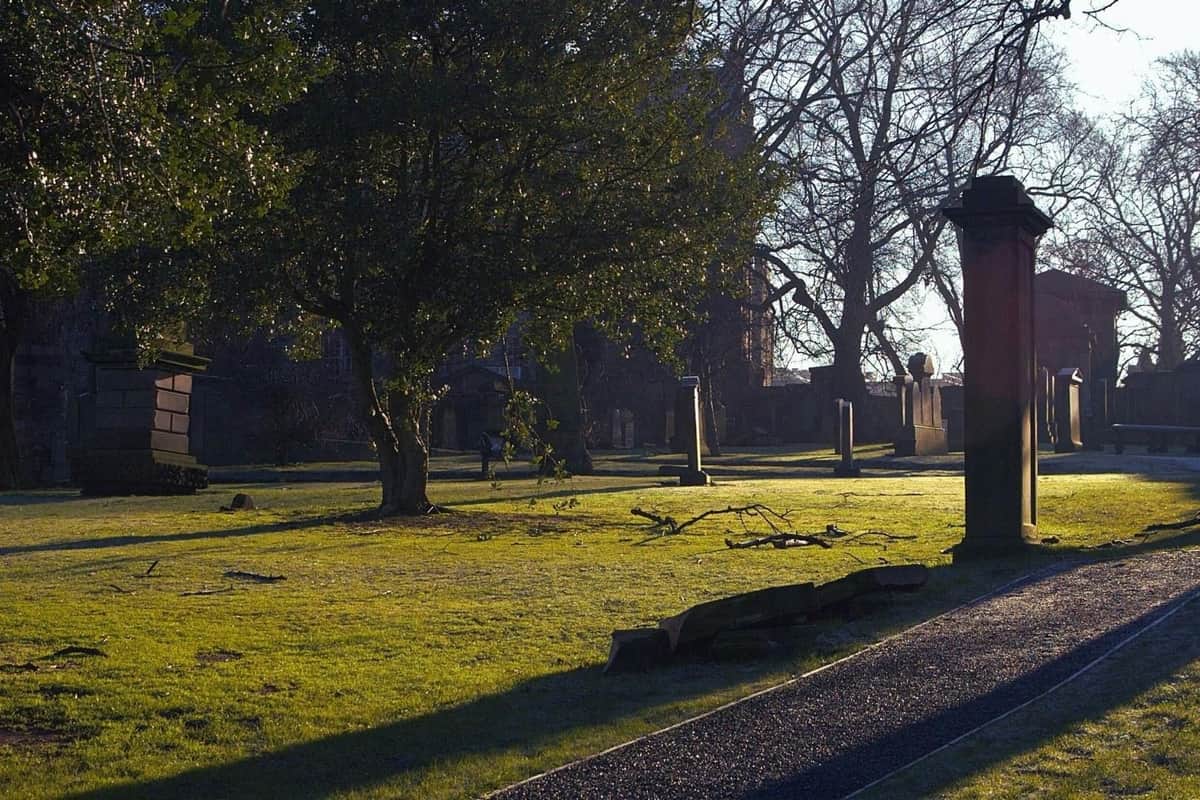 Edinburgh: 2-Hr Guided Ghost Tour A sunlit graveyard with trees, gravestones, and a pathway, showcasing a calm and serene atmosphere.
