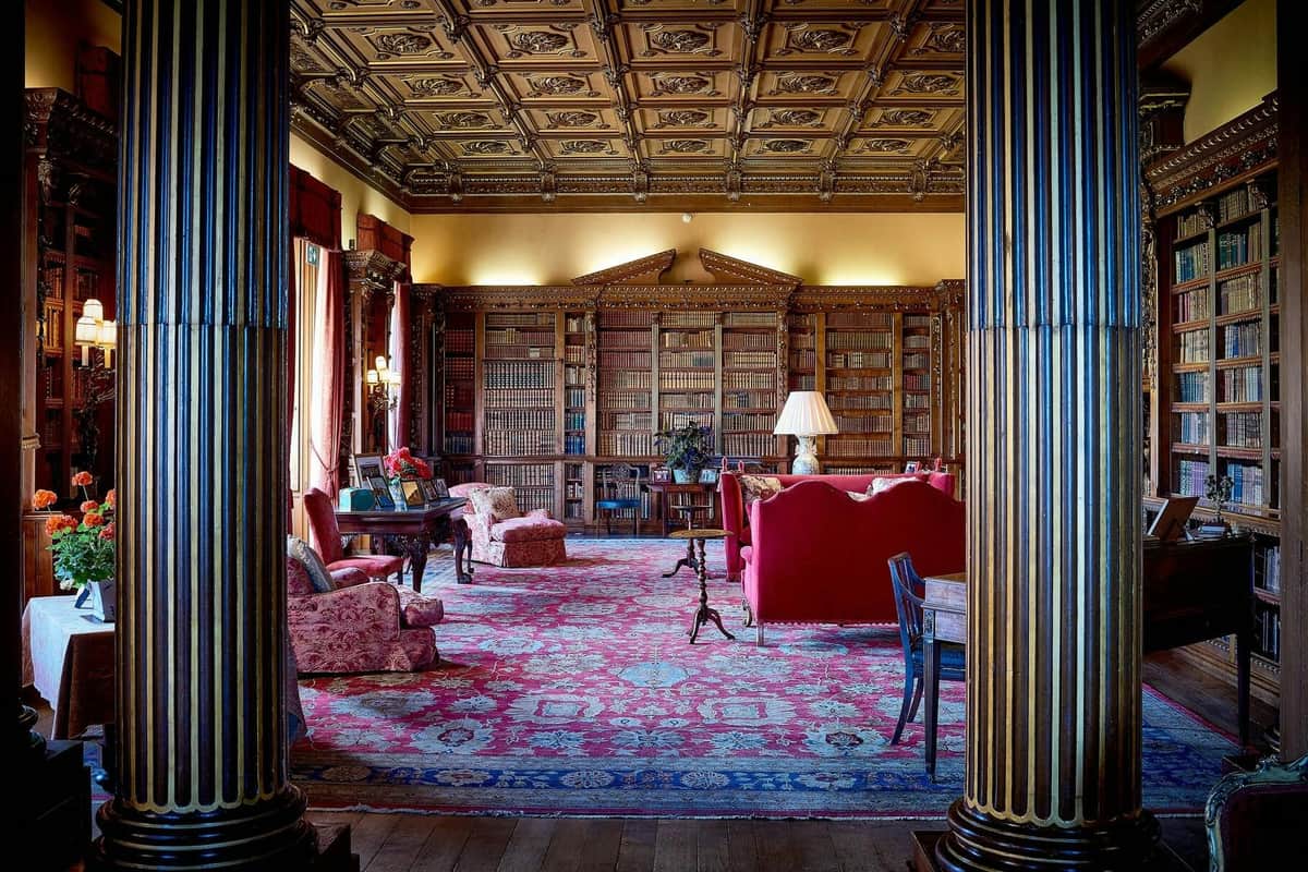 Une bibliothèque luxueuse avec des boiseries ornées, des étagères, des meubles rembourrés rouges et un plafond décoratif.