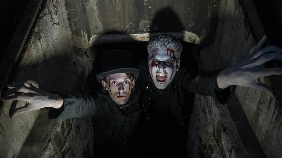 Shiverpool: Auld City & The Dead House Tour Two people with dramatic makeup and fake blood look upwards from a dark, enclosed space; one wears a top hat.