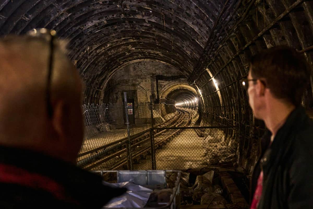 Holborn Londres : Visite guidée des stations de métro cachées Deux personnes observent un tunnel souterrain avec des voies ferrées, entouré de murs et éclairé par un éclairage industriel.