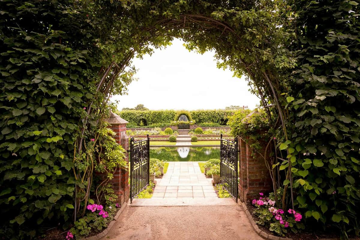 Un portail en fer forgé s'ouvre sur un chemin en briques menant à un étang réfléchissant, avec des jardins manucurés, des fleurs et un treillis arqué.