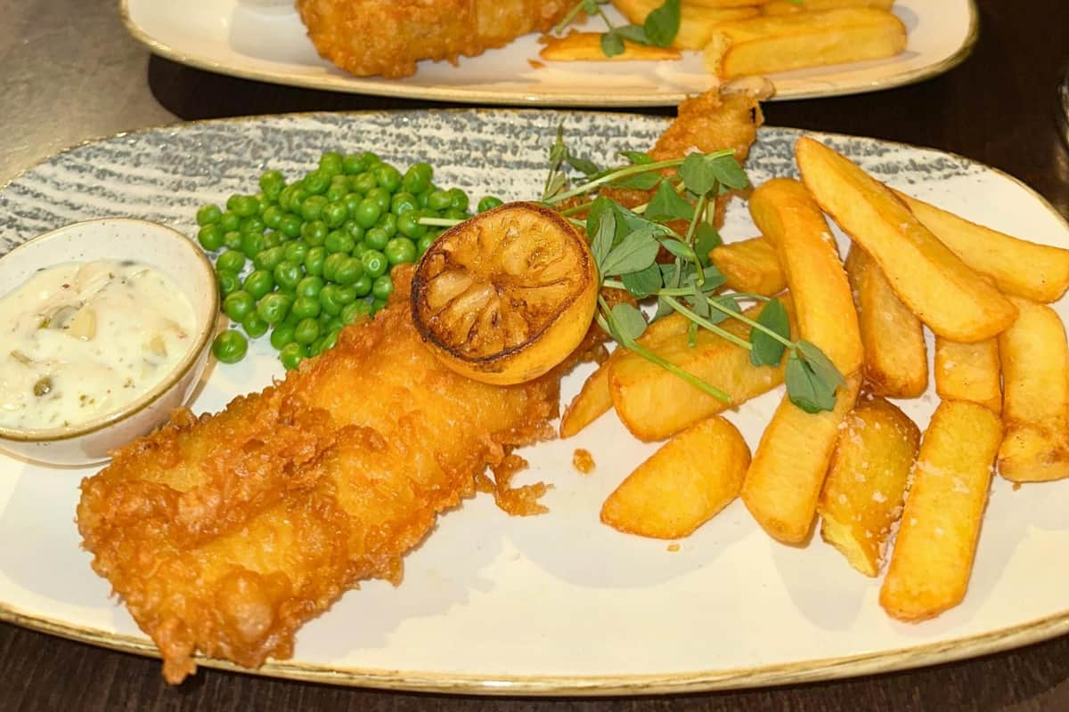 Londres : chasse au trésor gastronomique en autonomie, visite à pied et déjeuner dans un pub Une assiette contenant du poisson pané, une rondelle de citron, des petits pois, des frites et une garniture, le tout présenté dans un plat en céramique clair.