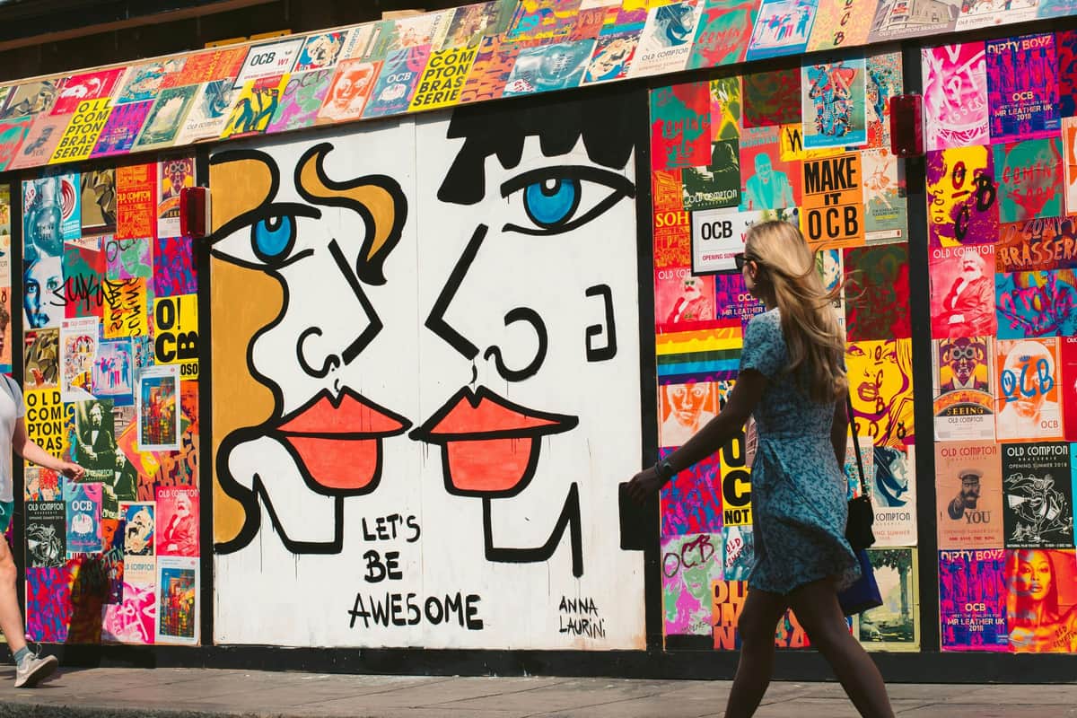 Londres Shoreditch : Visite à pied autoguidée en podcast Peinture murale de rue colorée représentant des visages abstraits avec des lignes audacieuses et des couleurs vives. Une personne vêtue d'une robe bleue passe devant.