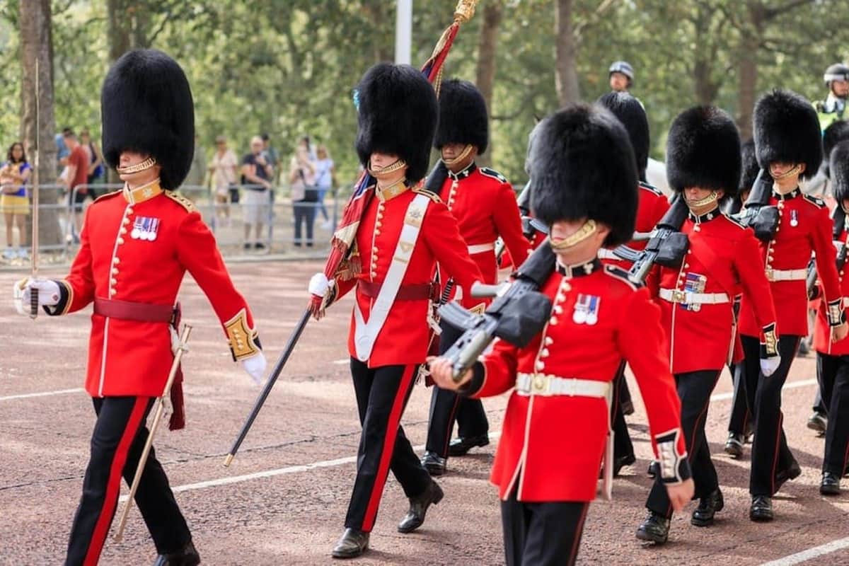 Londres : Visite privée à pied de Westminster + relève de la garde + prise en charge à l'hôtel Des gardes en uniformes rouges et grands chapeaux noirs marchent en formation, chacun tenant un fusil, sur fond de forêt.