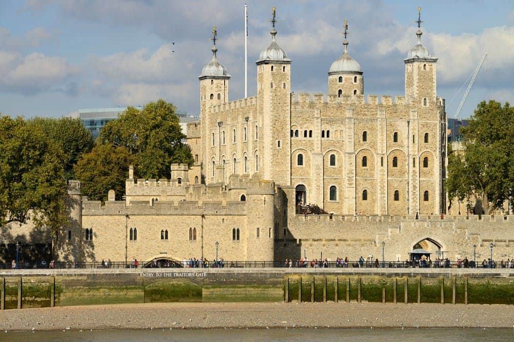 Tower of London: Early Guided Tour + Crown Jewels & Opening Ceremony Historic stone castle with towers and battlements, seen from across a river. Group of people walking near the entrance.