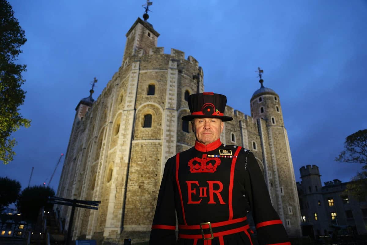 A Beefeater in ceremonial uniform stands in front of the illuminated Tower of London at dusk.