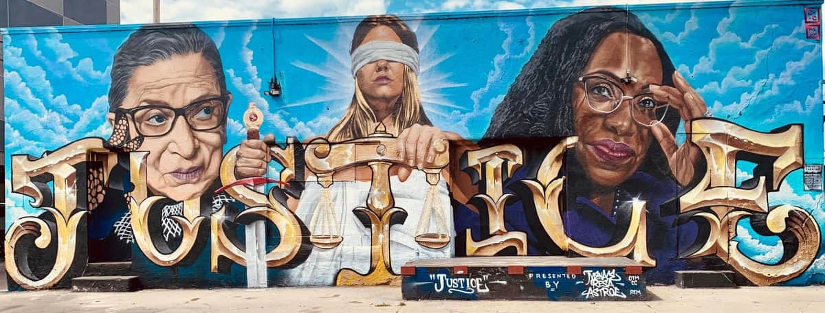 Denver Graffiti Original Tour (Denver) A vibrant mural depicting Lady Justice holding scales and a sword, with bold lettering and a blue sky background.