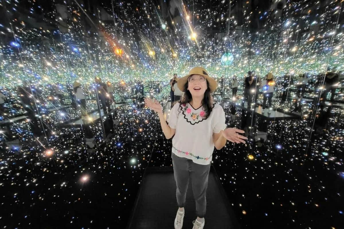 Los Angeles : Visite guidée de Downtown Art & Music (Los Angeles) Une femme se tient dans une pièce aux murs miroirs et aux lumières scintillantes. Elle porte un chapeau, des lunettes, un haut fleuri, un pantalon gris et des baskets.