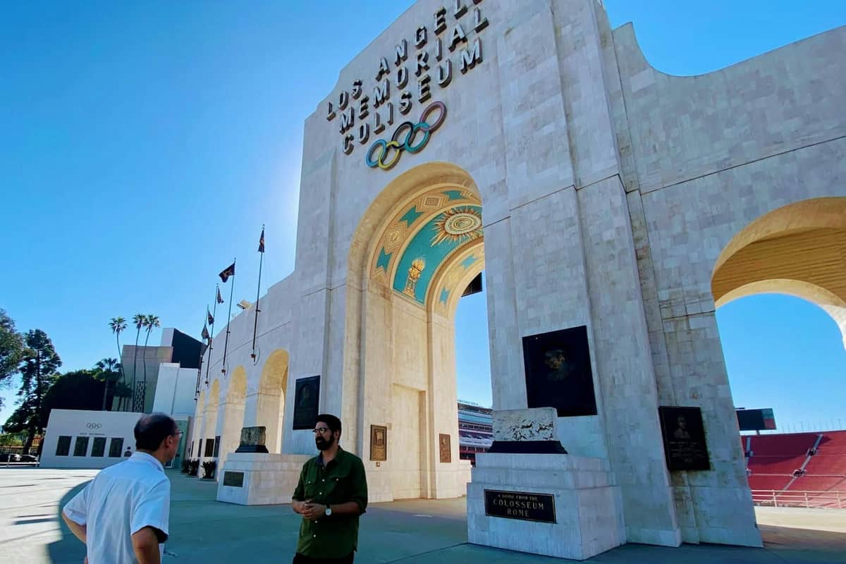 Deux personnes se tiennent à l'extérieur du Los Angeles Memorial Coliseum, avec ses anneaux olympiques et ses grandes arches, par temps clair.