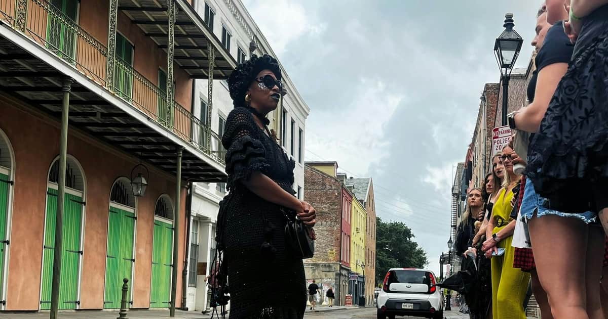 La Nouvelle-Orléans : visite guidée à pied sur le thème des fantômes, du vaudou et des vampires Une personne vêtue de noir se tient dans une rue, sous le regard d'un groupe de passants, entourée de bâtiments colorés et d'un ciel nuageux.