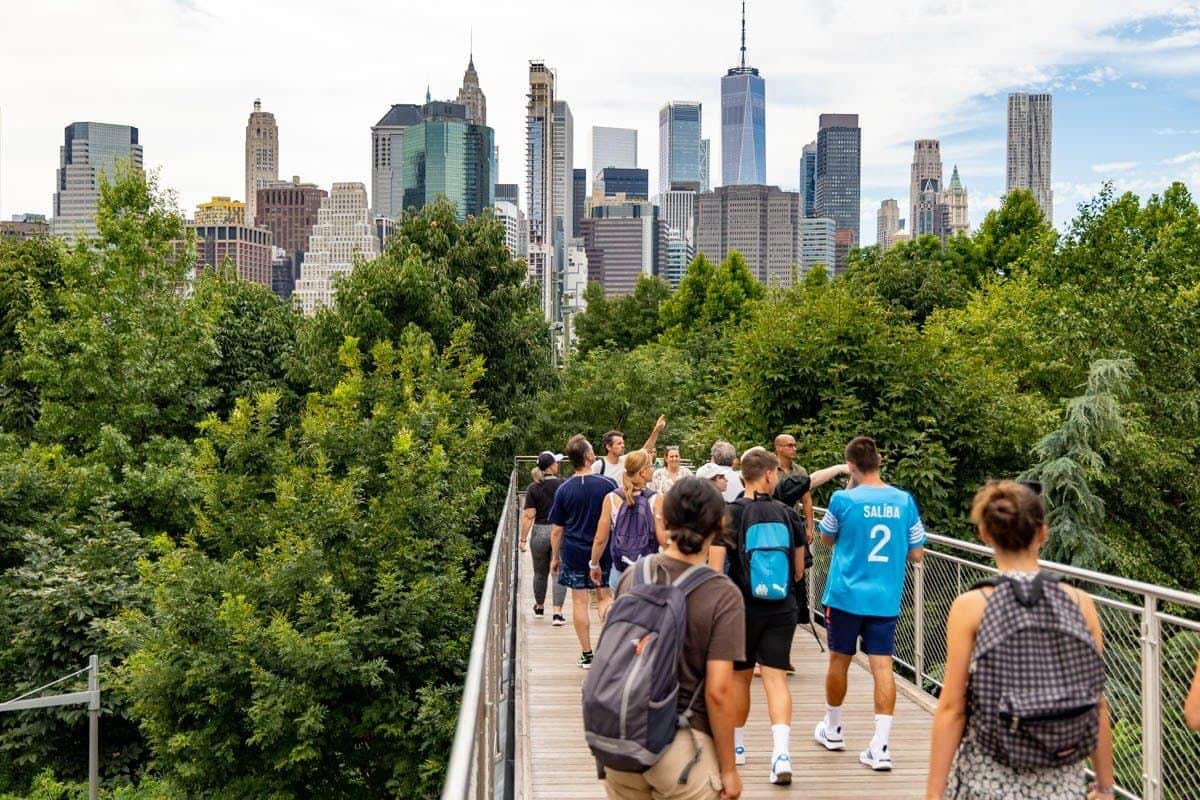 People walking on an elevated pathway surrounded by greenery, with a city skyline featuring skyscrapers in the background.