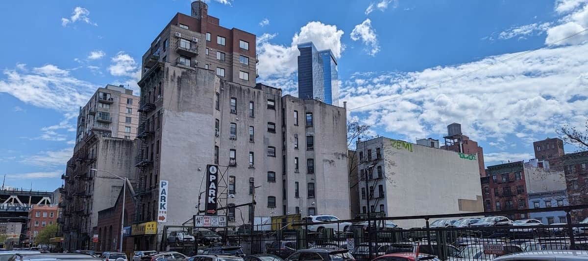 Visite guidée sur l'histoire des immeubles collectifs et des immigrés du Lower East Side Scène urbaine avec des voitures garées, des bâtiments anciens, une tour et un ciel partiellement nuageux.