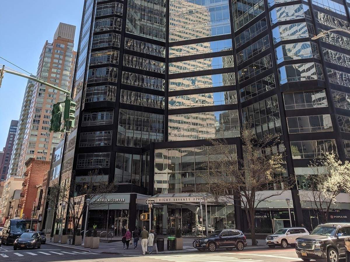 Les derniers résistants de Midtown : visite guidée à pied Un immeuble moderne aux façades vitrées, avec un Starbucks au rez-de-chaussée et quelques piétons et voitures à l'extérieur par une journée ensoleillée.