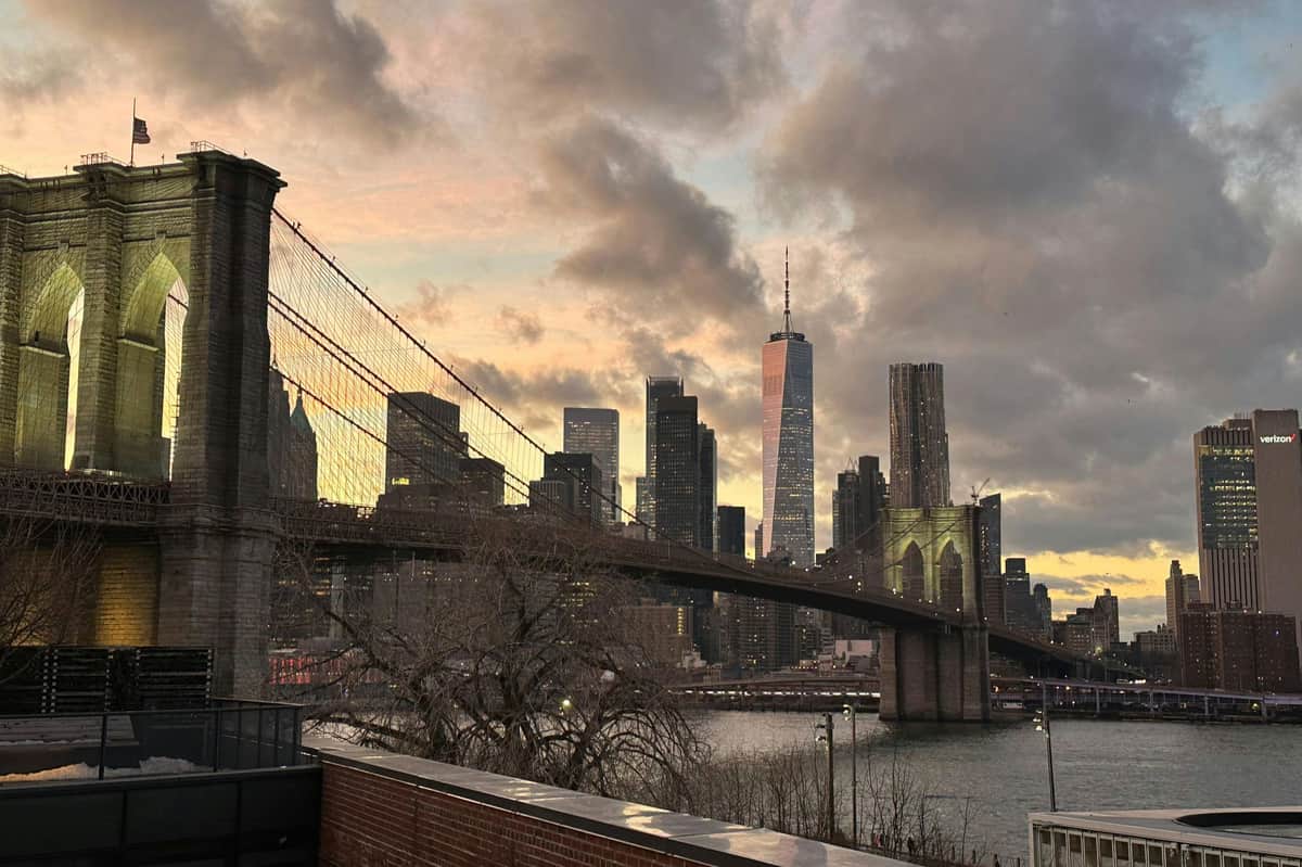 New York : Brooklyn Bridge & DUMBO Visite en espagnol Pont de Brooklyn au crépuscule avec l'horizon de la ville de New York en arrière-plan, avec le One World Trade Center et un ciel nuageux.