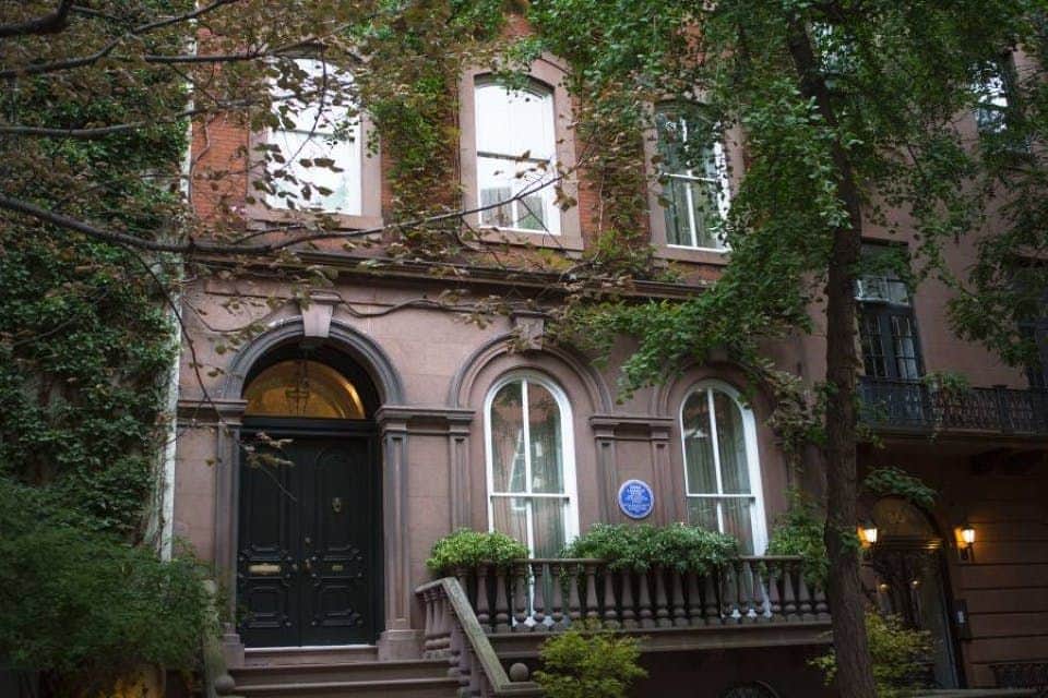 Three-story brownstone building with arched windows, black door, and a blue historical plaque, surrounded by green trees.