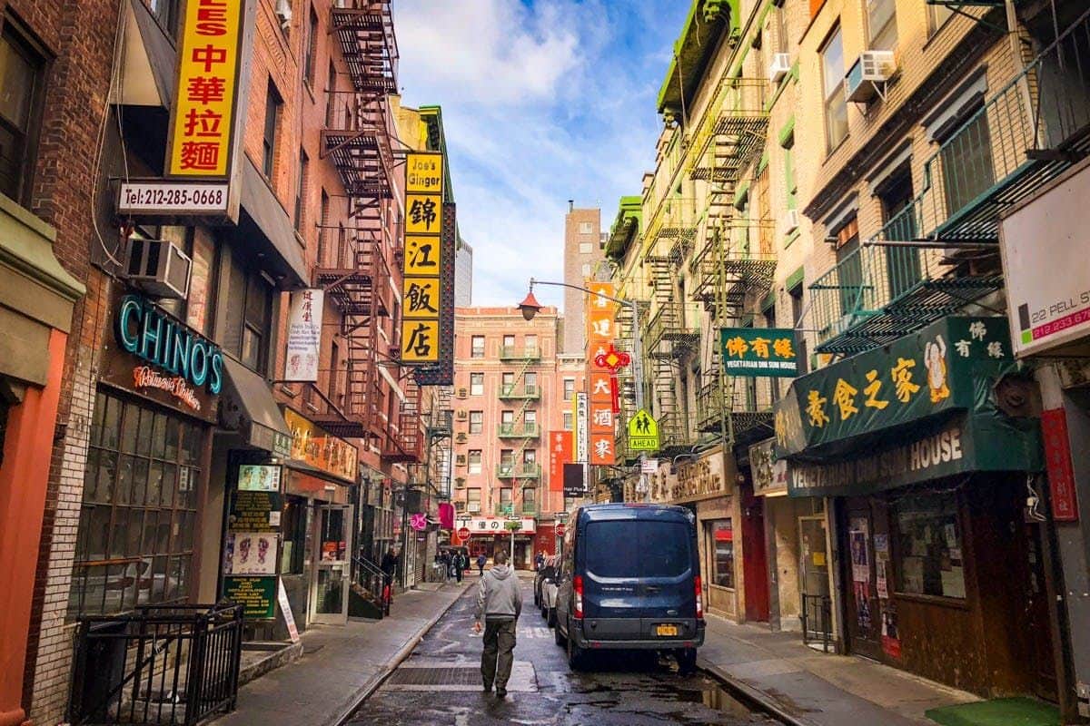 New York City : Lower East Side, Chinatown & Little Italy Visite en français Une rue étroite du quartier chinois avec des enseignes colorées, des escaliers de secours, des magasins, une camionnette bleue et une personne qui s'éloigne de l'appareil photo.