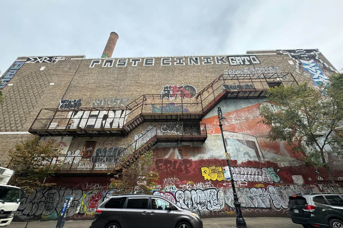 New York : Lower East Side Visite en espagnol Bâtiment en briques avec de nombreux graffitis, des escaliers de secours en métal et un monospace garé devant. Le ciel est couvert.