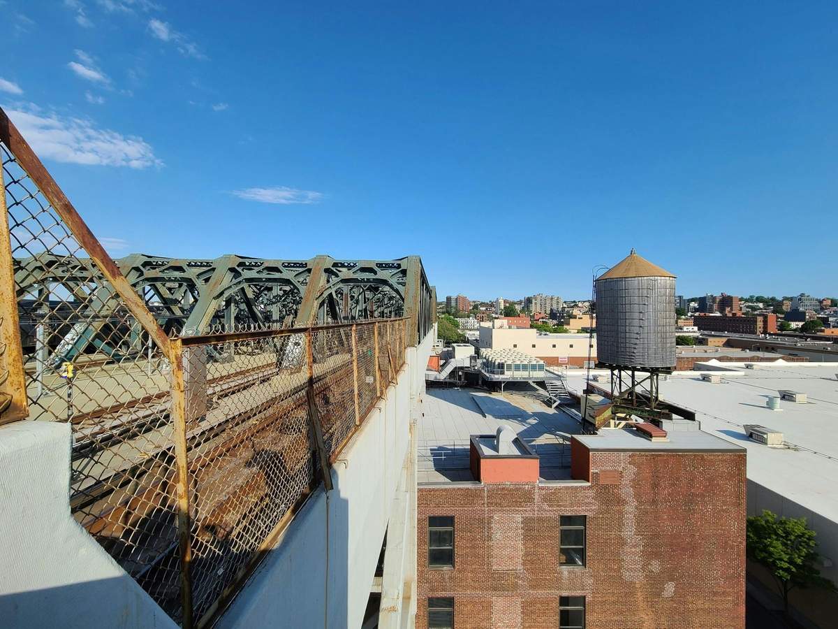 Visite guidée « Les secrets du métro de Brooklyn » Scène urbaine représentant un immeuble en briques, un château d'eau sur le toit et un pont métallique sous un ciel bleu limpide.