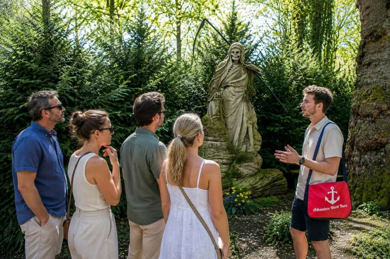 A tour guide is speaking to three visitors in front of a statue of a skeletal figure holding a scythe, set in a lush garden.