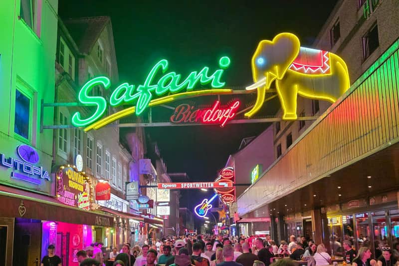 A bustling street at night with neon signs, including a large green and yellow one reading 'Safari Bierdorf,' and many people.